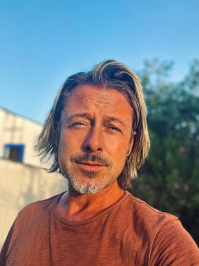 Man with half long blond hair and grey beard wearing brown t-shirt outside by spanish white house in countryside during golden hour.