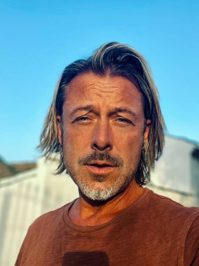 Man with half long blond hair and grey beard wearing brown t-shirt outside by spanish white house in countryside during golden hour.