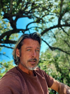 Man with half long blond hair and grey beard wearing brown t-shirt outside under a tree during summer.