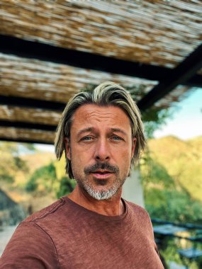 Man with half long blond hair and grey beard wearing brown t-shirt outside under a veranda in spanish countryside during summer.