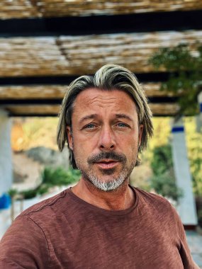 Man with half long blond hair and grey beard wearing brown t-shirt outside under a veranda in spanish countryside during summer.