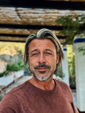 Man with half long blond hair and grey beard wearing brown t-shirt outside under a veranda in spanish countryside during summer.