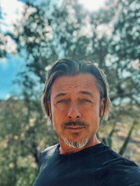 Sarı saçlı, sakallı, mavi tişörtlü, kır evindeki güneşli İspanyol bahçesinde oturan adam..