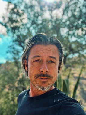 Sarı saçlı, sakallı, mavi tişörtlü, kır evindeki güneşli İspanyol bahçesinde oturan adam..