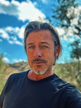 Sarı saçlı, sakallı, mavi tişörtlü, kır evindeki güneşli İspanyol bahçesinde oturan adam..