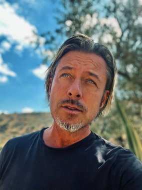 Sarı saçlı, sakallı, mavi tişörtlü, kır evindeki güneşli İspanyol bahçesinde oturan adam..