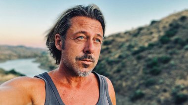 Orta yaşlı, bronz tenli, yarı sarı saçlı, gri sakallı, atlet gömlekli, altın saatinde dışarıda yürüyen bir adam. Almogia tepeleri, Malaga, Endülüs, İspanya.