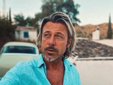 Bronz tenli, orta yaşlı, yarı sarı saçlı, mavi gömlekli, kırsal kesimdeki beyaz İspanyol evinde klasik arabalı bir adam. Malaga, Endülüs, İspanya 'da altın saat.