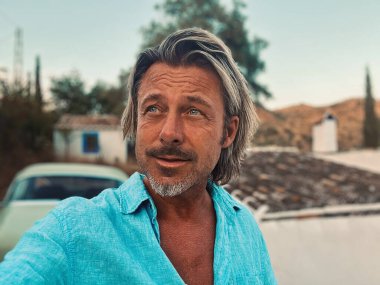 Bronz tenli, orta yaşlı, yarı sarı saçlı, mavi gömlekli, kırsal kesimdeki beyaz İspanyol evinde klasik arabalı bir adam. Malaga, Endülüs, İspanya 'da altın saat.