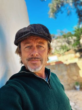 Yünlü gazeteci şapkası ve sakallı, yeşil yünlü süveteri olan haşin bir adam kırsal kesimdeki İspanyol evinin beyaz duvarının yanında duruyor..