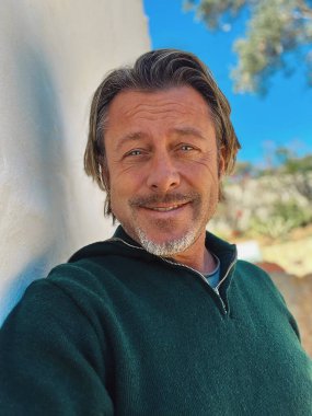 Yarı sarı saçlı, sakallı, yeşil yünlü kazaklı haşin bir adam. Kırsal kesimdeki İspanyol evinin beyaz duvarının yanında duruyor..