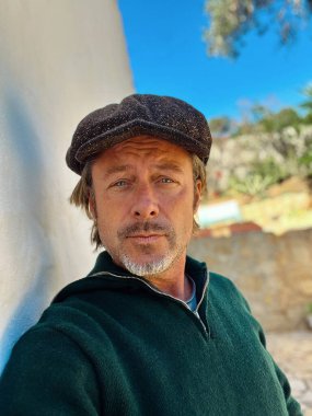 Yünlü gazeteci şapkası ve sakallı, yeşil yünlü süveteri olan haşin bir adam kırsal kesimdeki İspanyol evinin beyaz duvarının yanında duruyor..