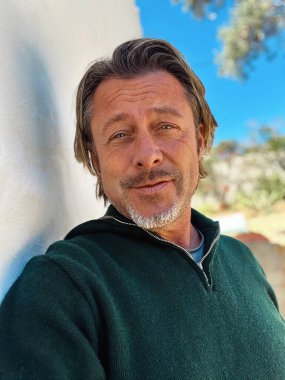 Yarı sarı saçlı, sakallı, yeşil yünlü kazaklı haşin bir adam. Kırsal kesimdeki İspanyol evinin beyaz duvarının yanında duruyor..