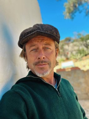 Yünlü gazeteci şapkası ve sakallı, yeşil yünlü süveteri olan haşin bir adam kırsal kesimdeki İspanyol evinin beyaz duvarının yanında duruyor..