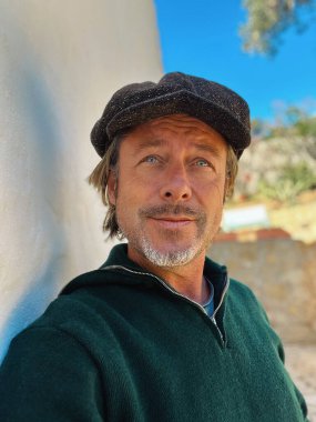 Yünlü gazeteci şapkası ve sakallı, yeşil yünlü süveteri olan haşin bir adam kırsal kesimdeki İspanyol evinin beyaz duvarının yanında duruyor..