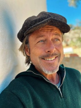 Yünlü gazeteci şapkası ve sakallı, yeşil yünlü süveteri olan, güler yüzlü haşin adam İspanyol evinin beyaz duvarının yanında, kırsal kesimde duruyor..