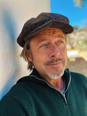 Yünlü gazeteci şapkası ve sakallı, yeşil yünlü süveteri olan haşin bir adam kırsal kesimdeki İspanyol evinin beyaz duvarının yanında duruyor..