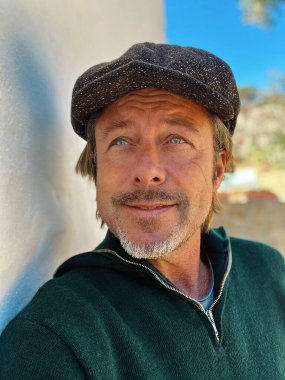 Yünlü gazeteci şapkası ve sakallı, yeşil yünlü süveteri olan haşin bir adam kırsal kesimdeki İspanyol evinin beyaz duvarının yanında duruyor..