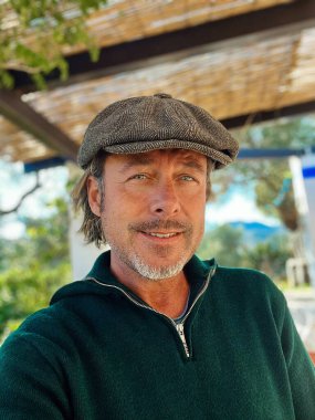 Yünlü gazeteci şapkası ve sakallı, yeşil yün süveterli, dışarıda verandanın altında mutlu, dayanıklı bir adam..