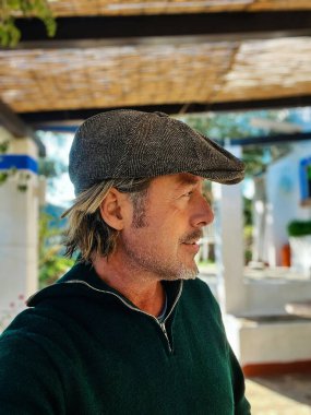 Yünlü gazeteci şapkası ve sakallı, yeşil yün kazaklı, verandanın altında haşin bir adam. Profil görünümü.