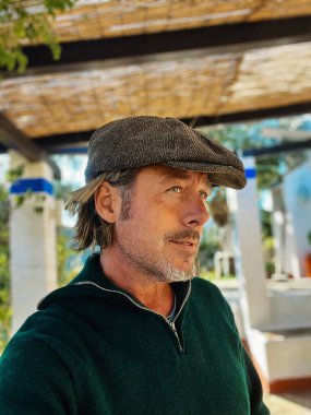 Yünlü gazeteci şapkası ve sakallı, yeşil yün kazaklı, verandanın altında haşin bir adam. Profil görünümü.