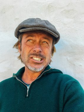 Yünlü gazeteci şapkası ve sakallı, yeşil yünlü süveteri olan etkileyici, haşin bir adam. İspanyol evinin beyaz duvarının yanında..