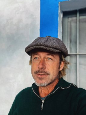 Yünlü gazeteci şapkası ve sakallı, yeşil yün kazağı olan haşin bir adam. İspanyol evi pencereli beyaz duvarın yanında..