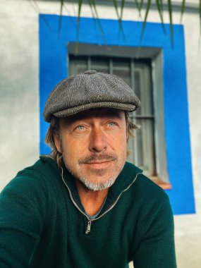 Yünlü gazeteci şapkası ve sakallı, yeşil yün kazağı olan haşin bir adam. İspanyol evi pencereli beyaz duvarın yanında..