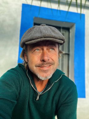 Gülen sert adam, yünlü gazeteci şapkası ve sakallı yeşil yün süveter giyen, İspanyol evi pencereli beyaz duvarlı..