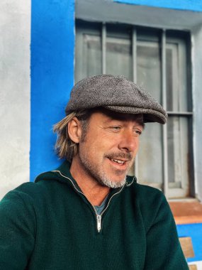 Gülen sert adam, yünlü gazeteci şapkası ve sakallı yeşil yün süveter giyen, İspanyol evi pencereli beyaz duvarlı..