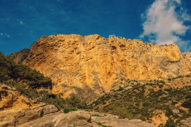 Dik, uzun, turuncu kayalık yamaçta mavi gökyüzünün altında bulutlu yeşil bir bitki örtüsü var. Caminito del Rey, Malaga, Endülüs, İspanya.