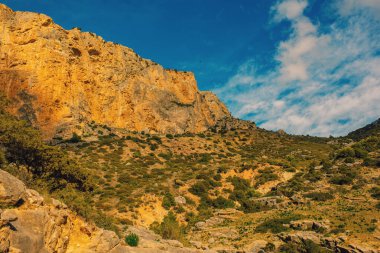 Pürüzsüz, uzun, turuncu kayalık yamaçlar. Mavi gökyüzünün altında, bulutlu yeşil bir bitki örtüsü. Caminito del Rey, Malaga, Endülüs, İspanya.