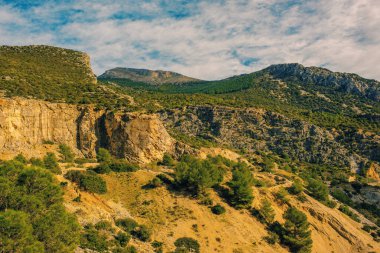 Sarp, turuncu kayalık dağlar, mavi gökyüzünün altında bazı yeşil bitkiler ve bulutlar. Sierra de Huma, Malaga, Endülüs, İspanya.
