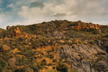 Bulutlu bir gökyüzünün altında bitki örtüsü, çalılık ve ağaçlarla kaplı sert kayalık bir tepe. Huebro, Almerya, Endülüs, İspanya.