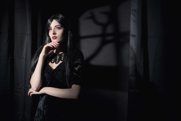 Dark mysterious witch fashion woman. Black curtains with window shadow on wall.