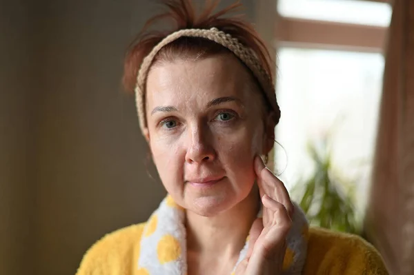 Portrait adult Caucasian woman applies cosmetic mask with fingers. The model stands at the window of the house. — Fotografia de Stock