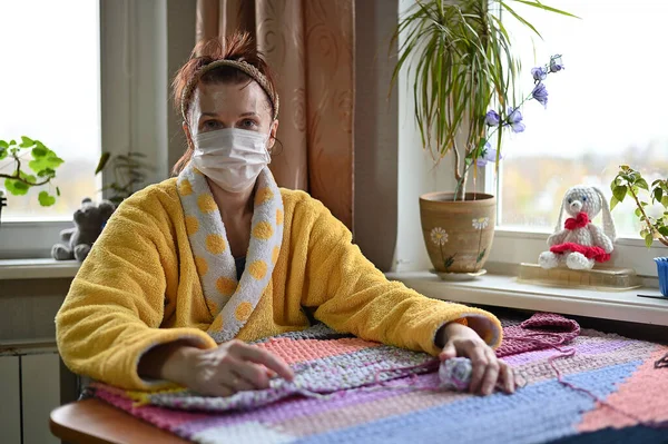 Portrait woman in a medical mask in a yellow coat at home knits with her hands. The model is engaged in needlework by the window. — Stock fotografie