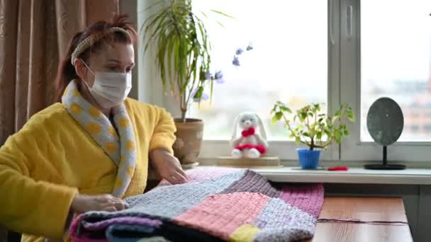Portrait of an aged woman 45 years old in a mask in quarantine at home knits with her hands while sitting at a table near the window — Stockvideo