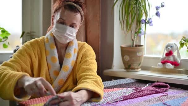 Портрет пожилой женщины 45 лет в маске в карантине дома вяжет руками, сидя за столом у окна — стоковое видео