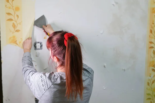 Una mujer adulta se quita el papel pintado trabaja con una espátula en la habitación. Concepto de renovación del hogar. —  Fotos de Stock