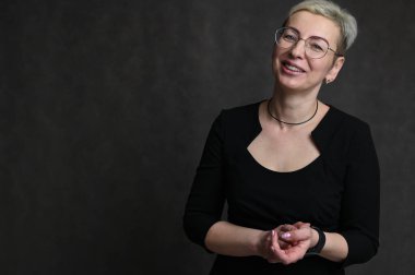 Siyah arka planda kısa saçlı, gözlüklü, sarışın, beyaz bir kadının fotoğrafı.