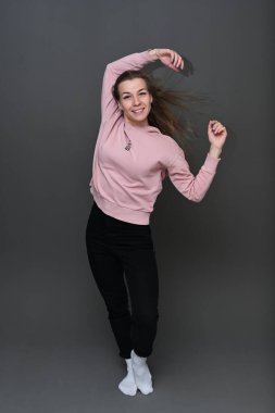 Portrait of a Caucasian girl with fluttering hair in the wind in black trousers and a pink sweater with a smile on a gray background in full growth. Studio photo in high resolution.