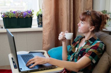 Kafkasyalı kadın, evinde, kanepede dizüstü bilgisayarla kahve içiyor.