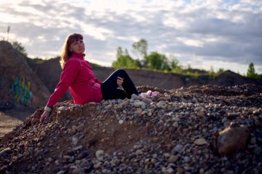 Gökyüzünün arka planında, kumların üzerinde otururken kameraya bakan ceketli beyaz bir kadının fotoğrafı.