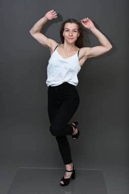 Beyaz bluzlu, siyah pantolonlu, gri arka planda gülümseyen, beyaz, beyaz bir kızın tam boy stüdyo resmi.