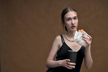 Kameraya kahve ve hamburgerle poz veren beyaz bir genç kız stüdyoda bej arka planda