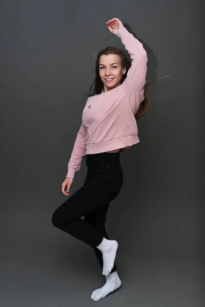 Full length photo of a happy young girl in a pink sweater and black trousers posing with flying hair on a gray background.