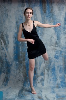 Young model in black dress in studio posing standing on one leg on blue background