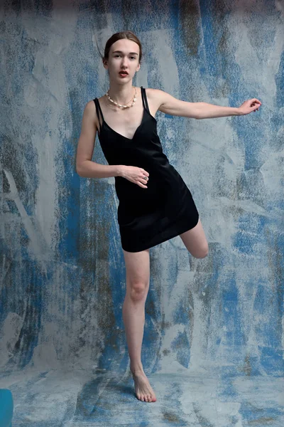 Young model in black dress in studio posing standing on one leg on blue background