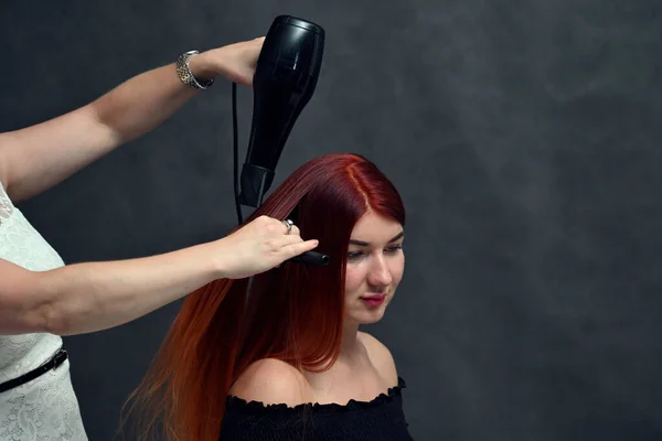 Hands of hairdresser doing hair with hairdryer of red-haired model on gray background